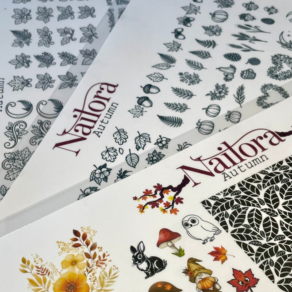 Three sheets of nail art DIY decals  featuring autumn-themed designs, including leaves, acorns, pumpkins, flowers, a rabbit, a mushroom, and an owl. The sheets are branded with 'Nailora Autumn' in a dark red font.