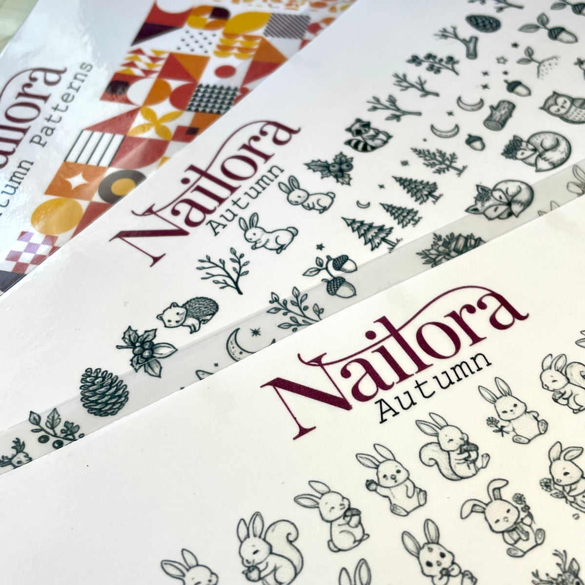 Three nail art pattern sheets from the brand Nailora, featuring autumn-themed designs like rabbits, squirrels, pinecones, and trees in black and gray line art on white backgrounds. Simple way to create your own nail art decals