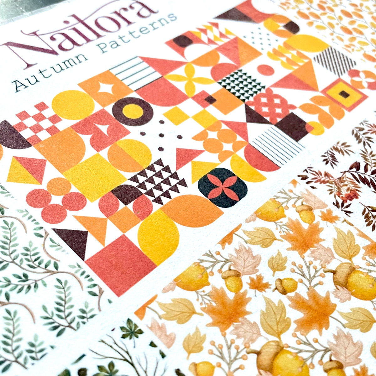 A close-up of a Nailora Autumn Patterns sheet featuring a colorful geometric design in orange, yellow, red, and brown, with another patterned sheet showing autumn leaves and acorns.